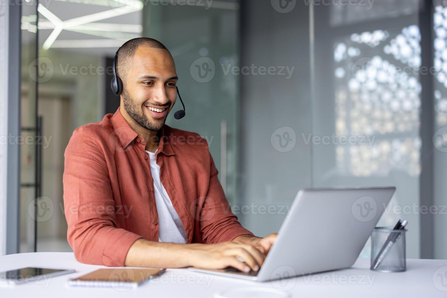 Smiling professional wearing headset works on laptop in modern office setting. Engaged in online meeting using phone technology. Notebook and pen on table indicate readiness and focus. photo
