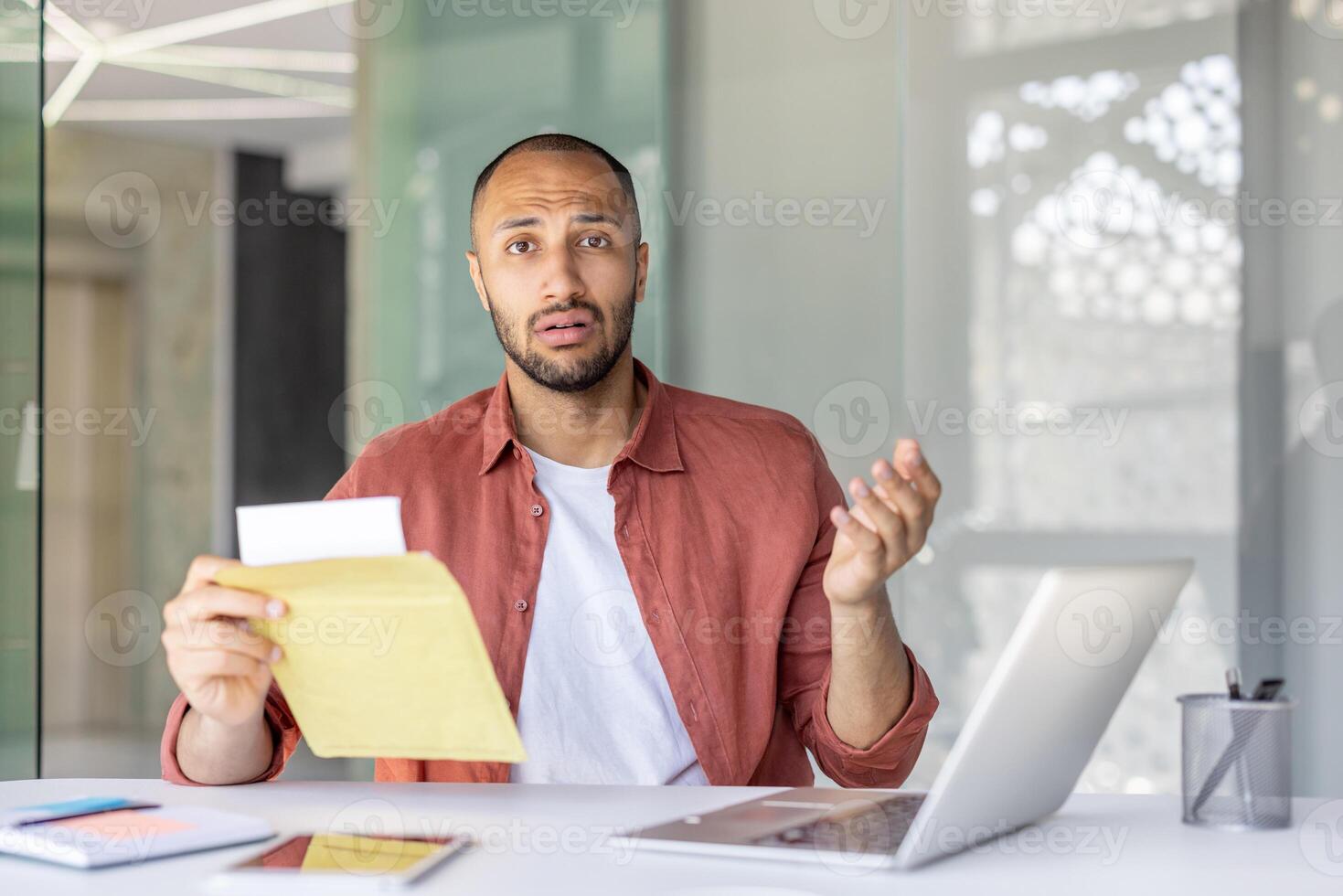 A confused office worker holds an envelope while sitting at a desk with a laptop. His expression suggests surprise or misunderstanding. Various devices and papers are on the desk. photo