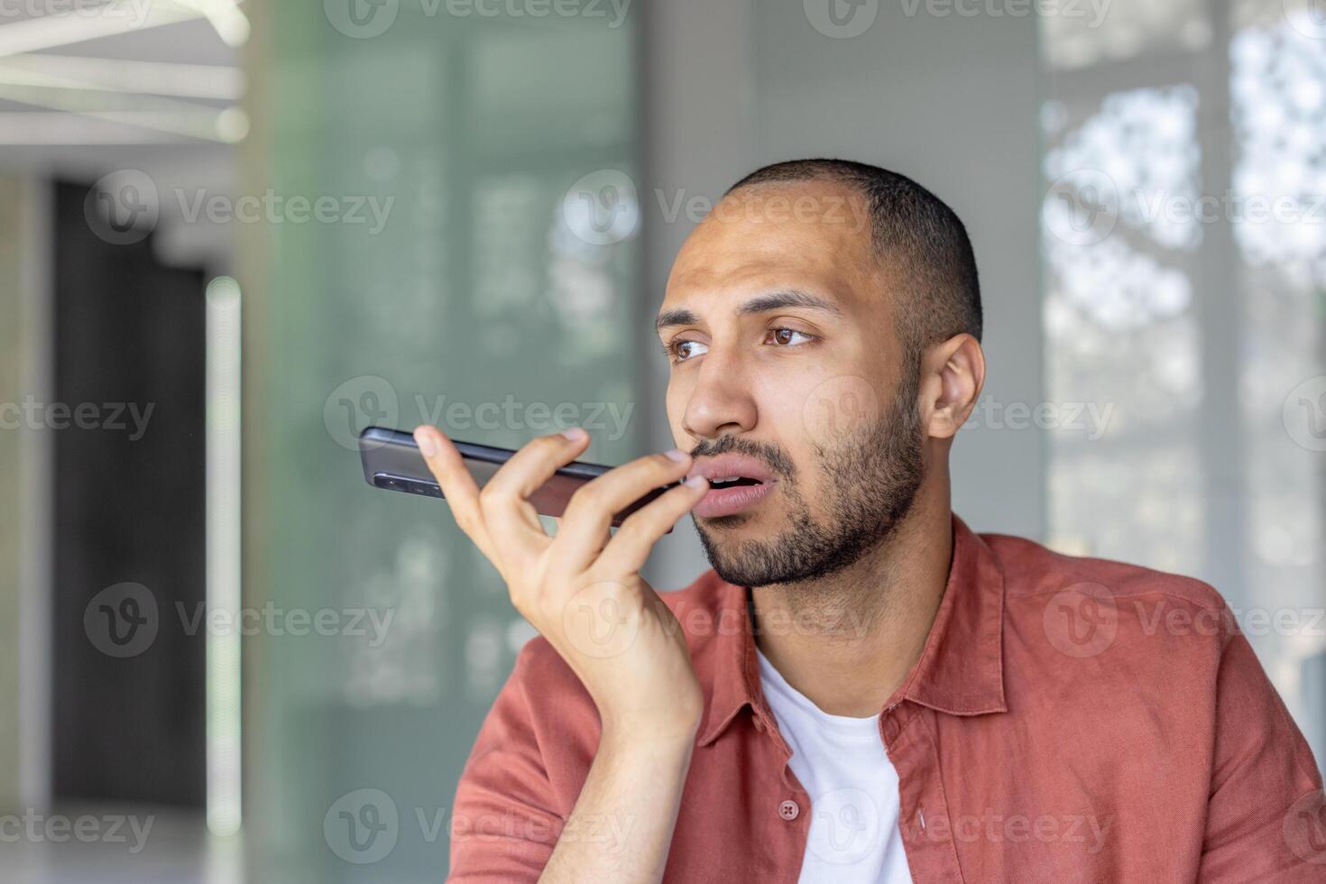 A man speaking into his smartphone, appearing engaged, likely recording or sending a voice message. The setting is indoors with a modern and professional atmosphere photo