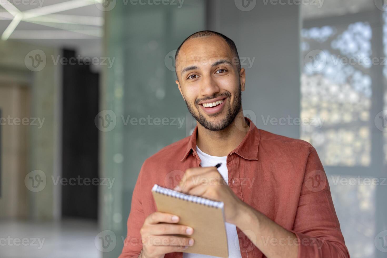 A confident man with a friendly smile is holding a notepad and pen inside a modern space, showcasing a positive and productive atmosphere. photo