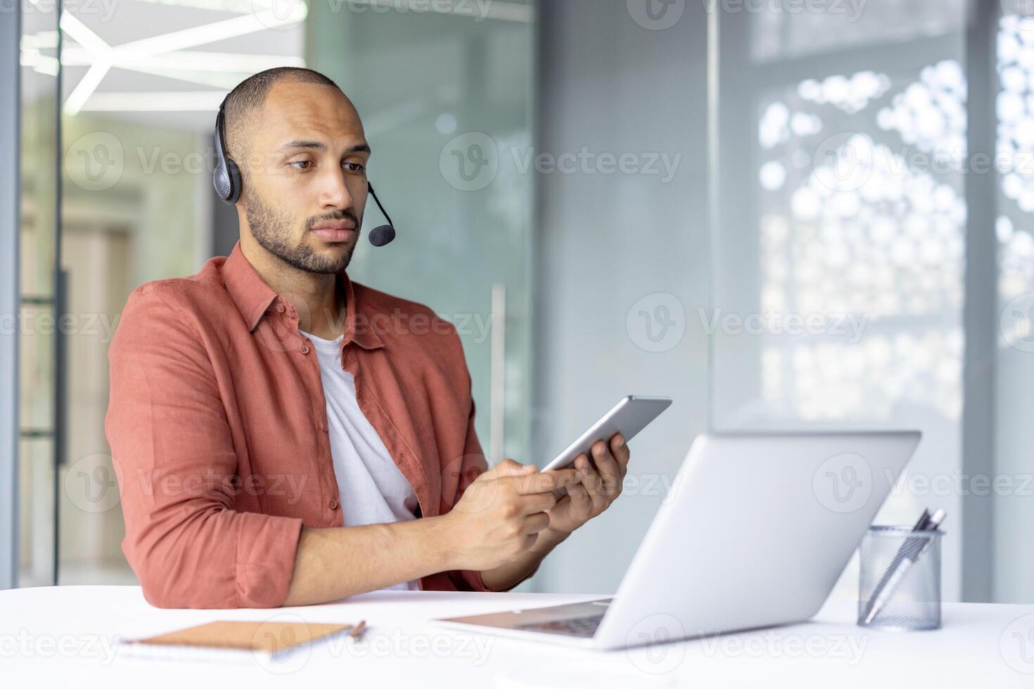 A young professional man is using a smartphone and laptop at his desk, wearing a headset. The scene suggests a remote work environment, highlighting technology use and communication. photo