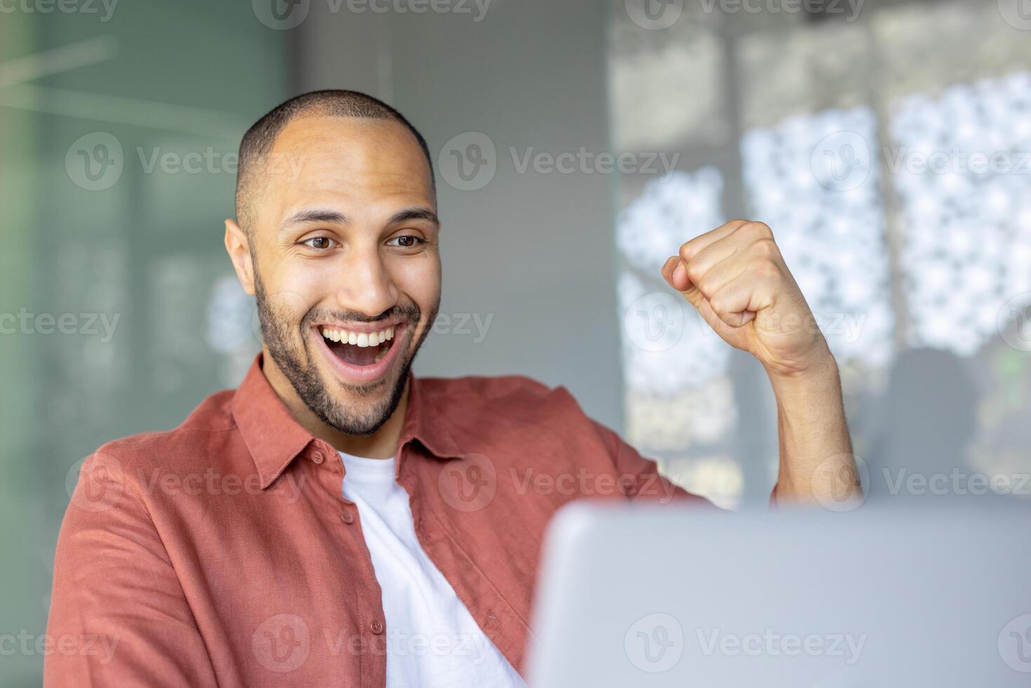 Smiling young man with joyful expression celebrates success while working on his laptop in an office environment. Concept of achievement, happiness, and professional success in a modern workspace. photo