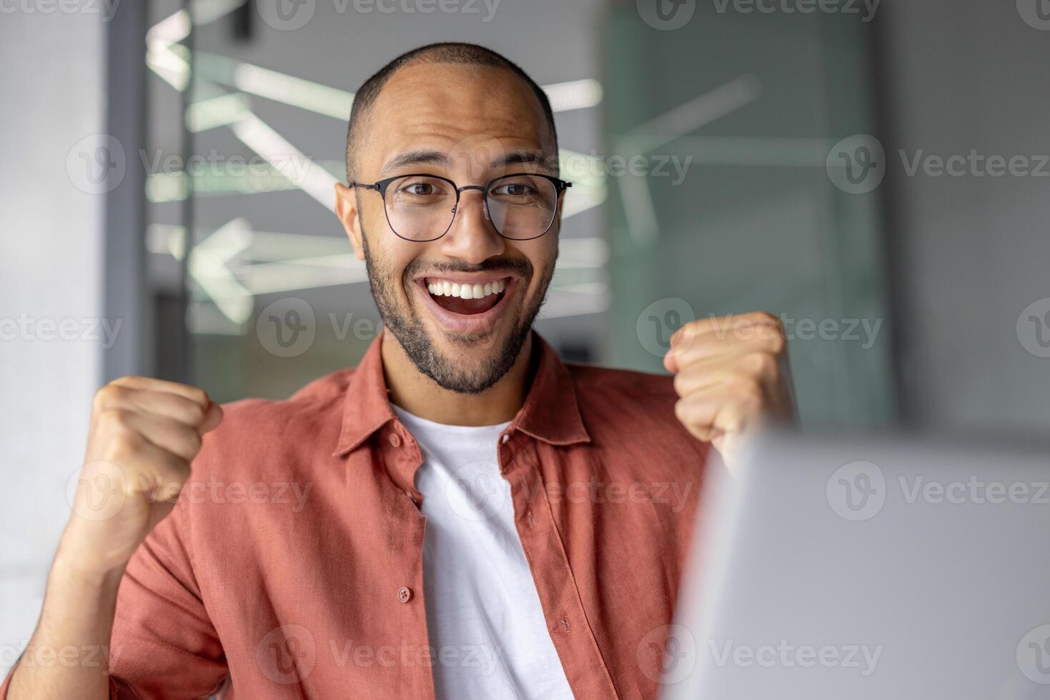 Energetic young man with glasses celebrating success with fists raised and joyful expression in modern office environment, conveying motivation and achievement. photo