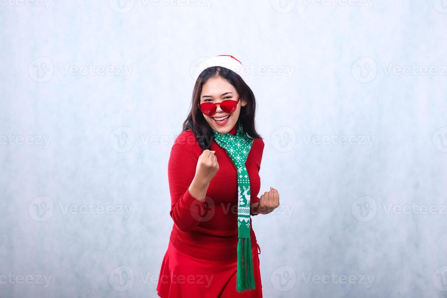 expression of asian woman wearing red sweater with Santa hat and scarf celebrating merry christmas, happy spirit to camera wearing glasses both hands clenched up and down, isolated on white background photo