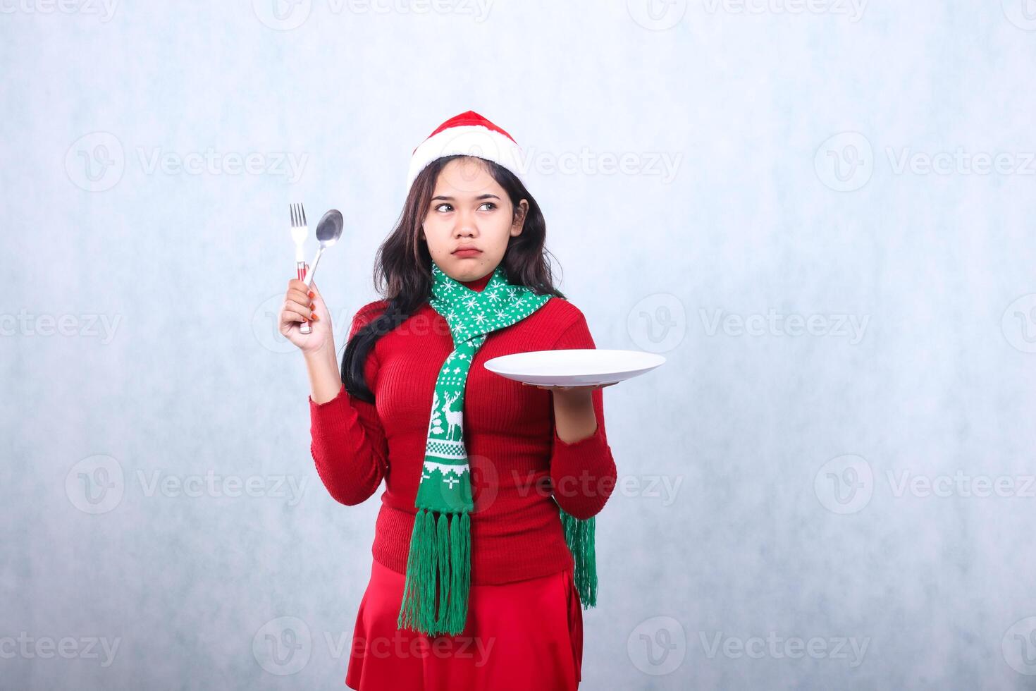expression woman wearing red sweater red sweater with santa hat and scarf celebrating merry christmas, annoyed to the left carrying white plate and holding spoon fork, isolated on white background photo