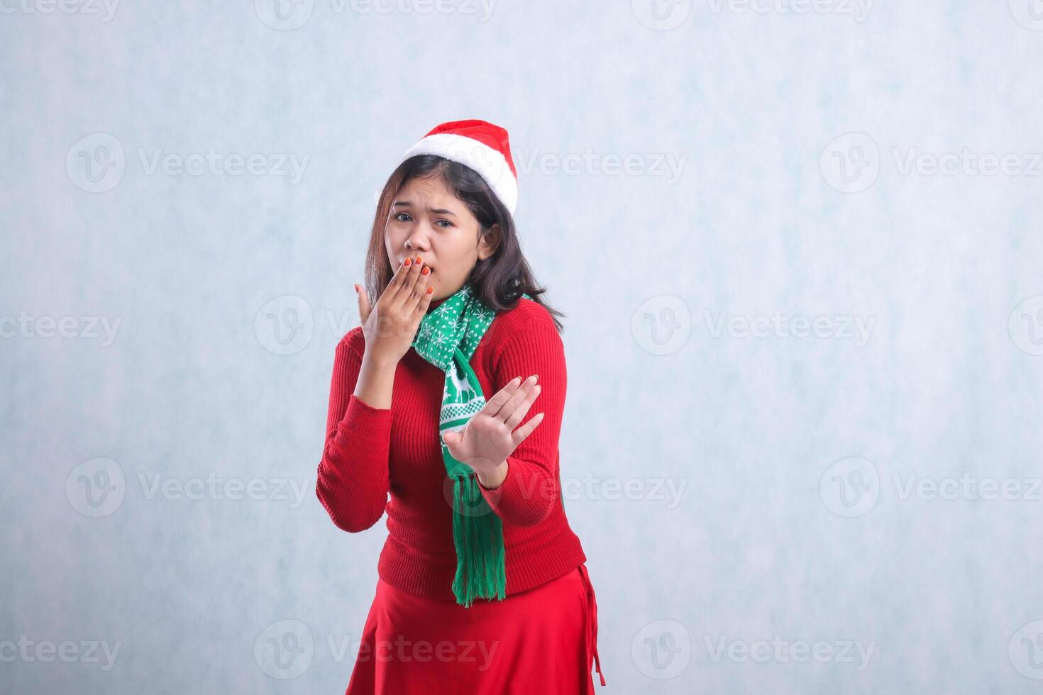 portrait of beautiful female wearing christmas sweater, santa hat and scarf, afraid to camera, hand sign reject to camera, covering mouth, isolated on white background photo