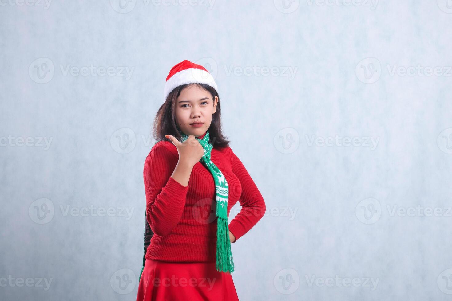 expression of young indonesia woman wearing christmas sweater, santa hat and scarf, slanting left pouting to camera hand sign pointing to right, isolated on white background photo