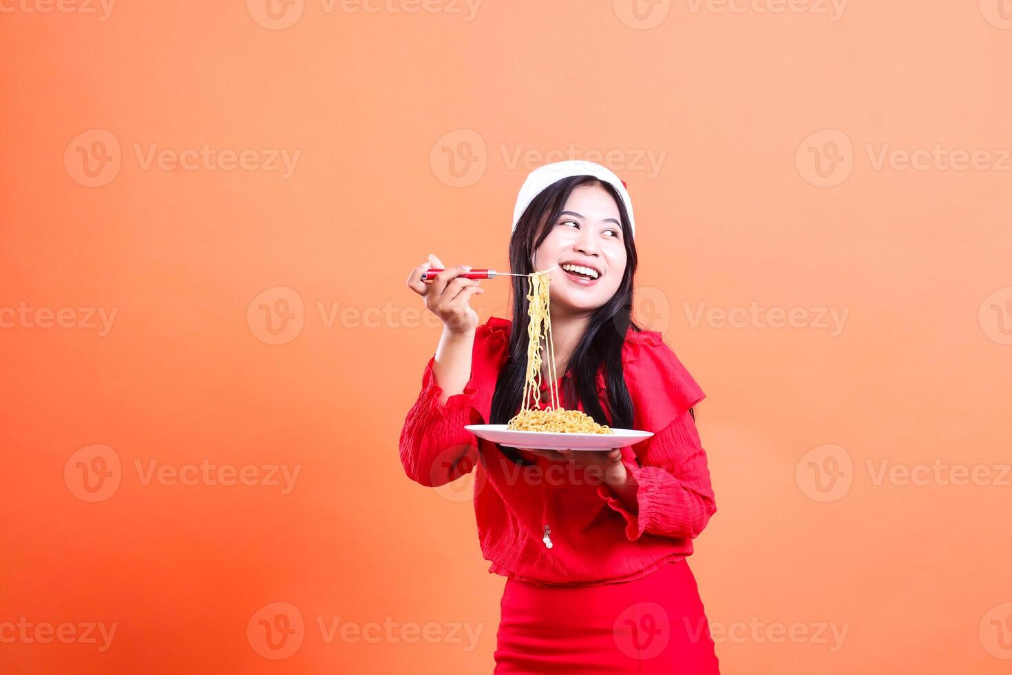 Asian ladies beauty laughing to left, hand holding fork eating noodles from white plate celebrating christmas, wearing red christmas blouse, isolated on orange background photo
