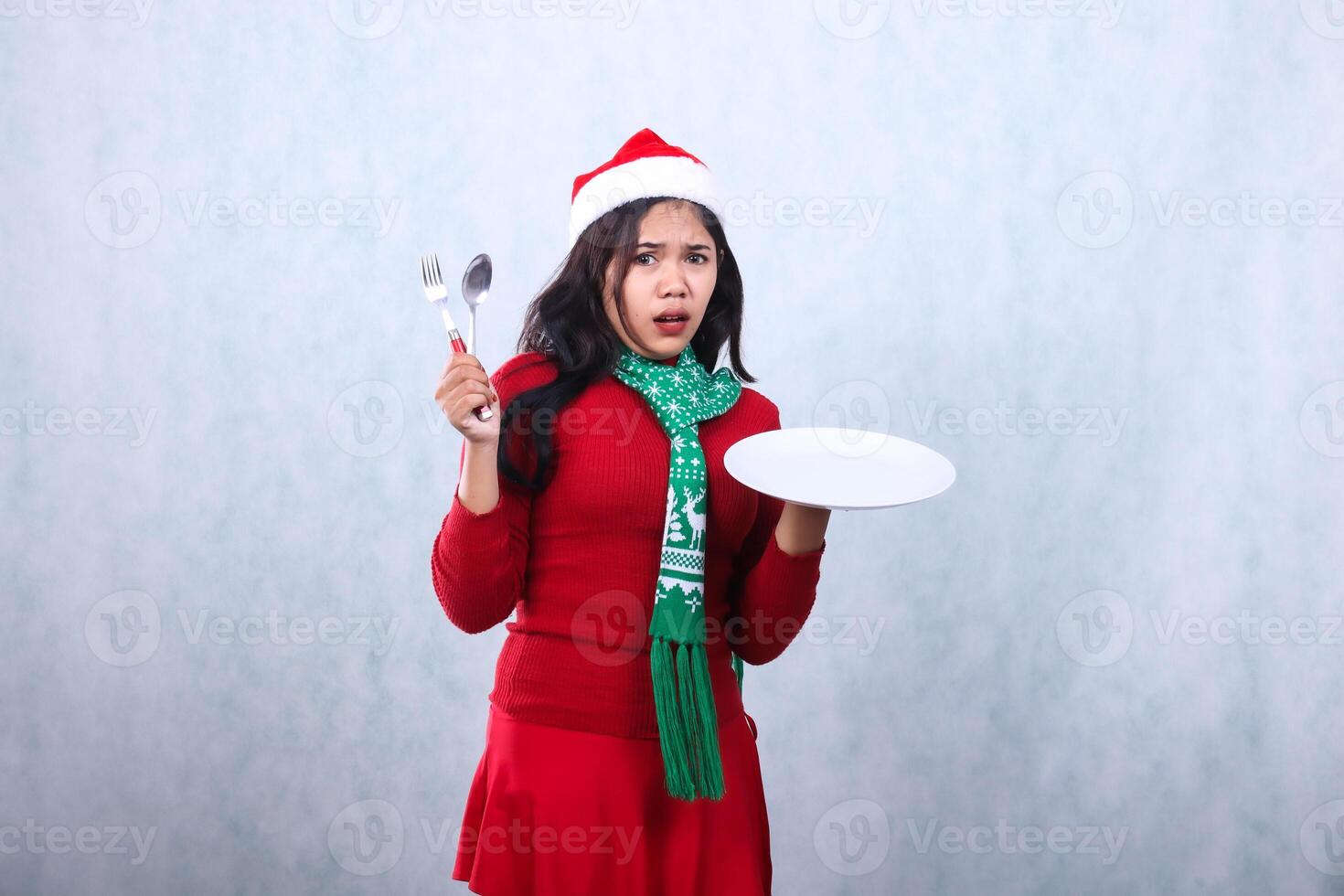 beautiful woman wearing red sweater red sweater with santa hat and scarf celebrating merry christmas, surprised to camera carrying white plate and holding fork spoon, isolated on white background photo