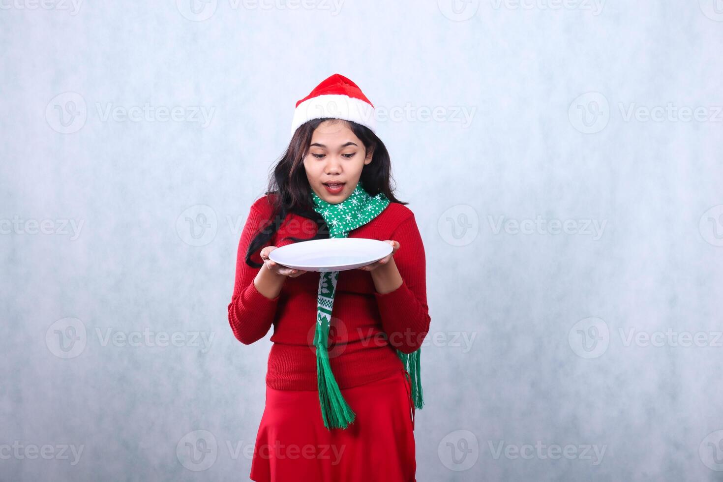 asian girl wearing red sweater with Santa hat and scarf, amazed looking at plate both hands holding white plate, celebrating merry christmas and new year isolated on white background photo