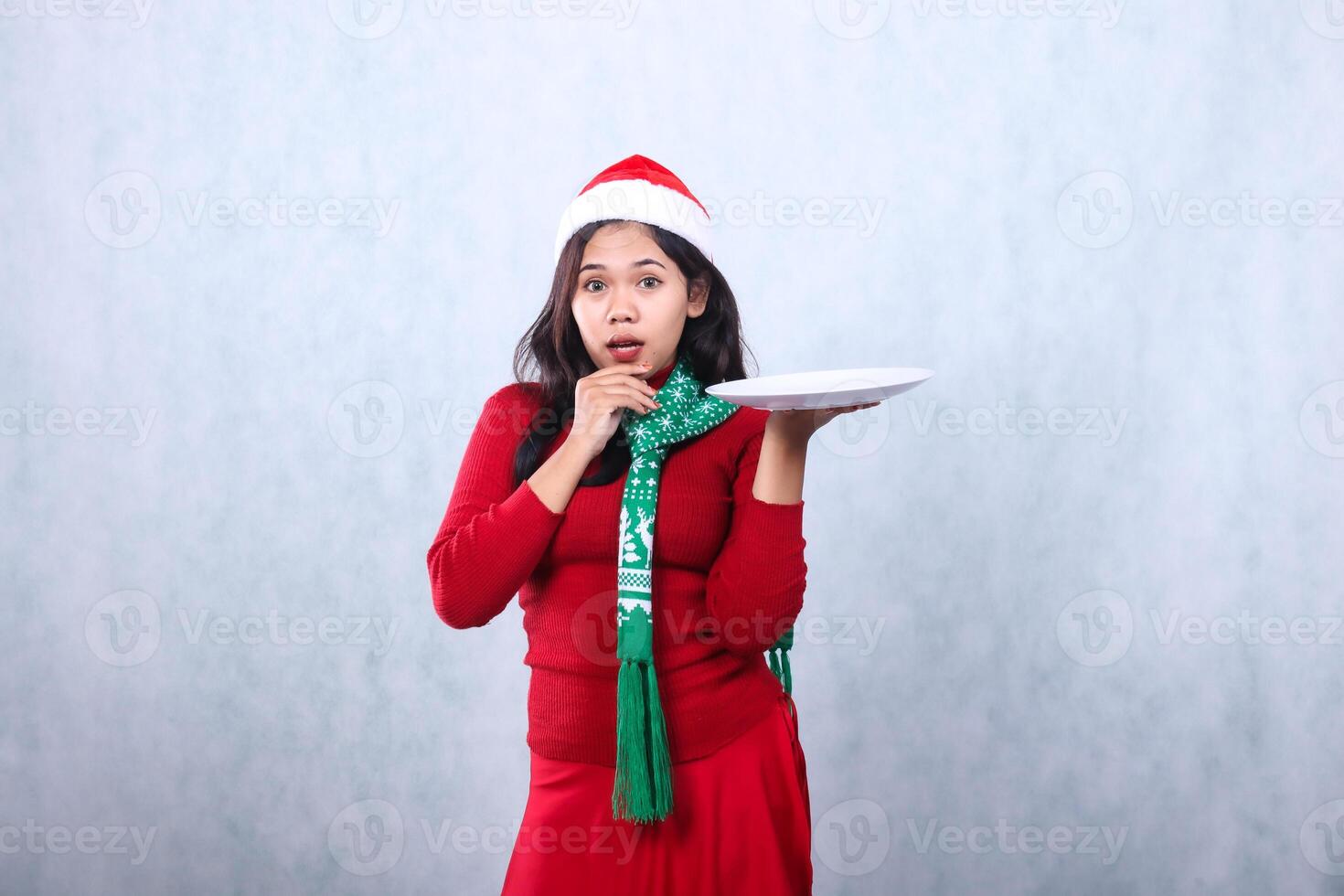 portrait of woman wearing red sweater red sweater with santa hat and scarf celebrating merry christmas, shocked looking at camera hand lift white plate and hold chin, isolated on white background photo