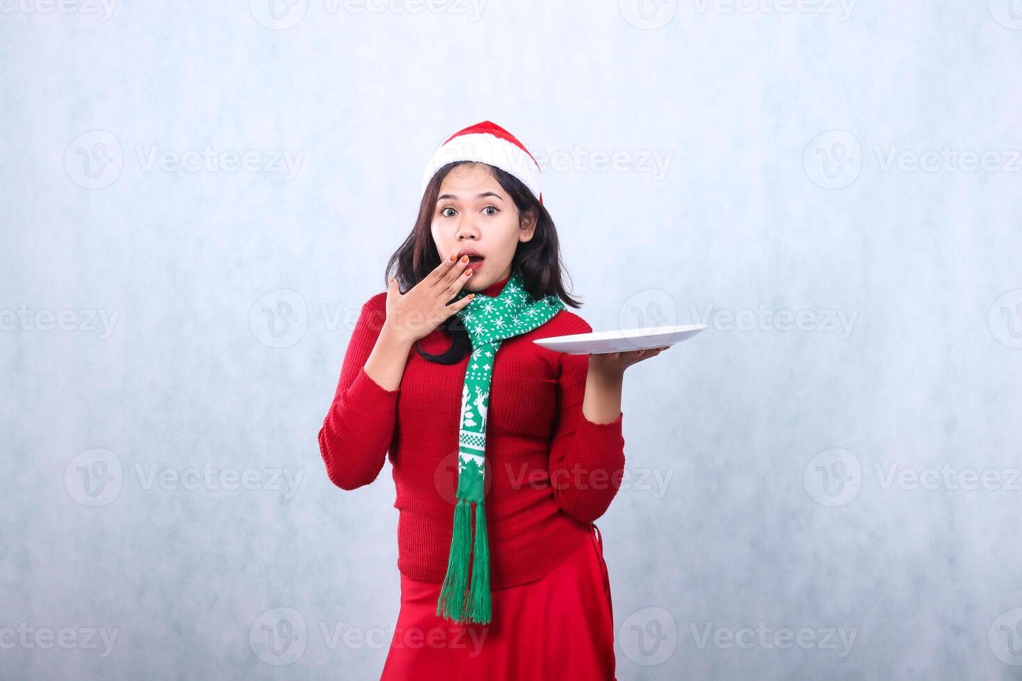 woman wearing red sweater with Santa hat and scarf, shocked looking at camera, hand lifting white plate and covering mouth, celebrating merry christmas and happy new year isolated on white background photo