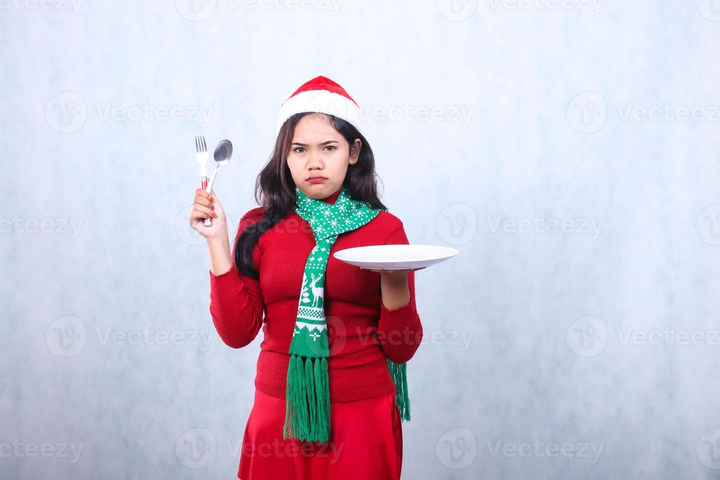 portrait asian woman wearing red sweater red sweater with santa hat and scarf celebrating merry christmas, annoyed to camera carrying white plate and holding spoon fork, isolated on white background photo