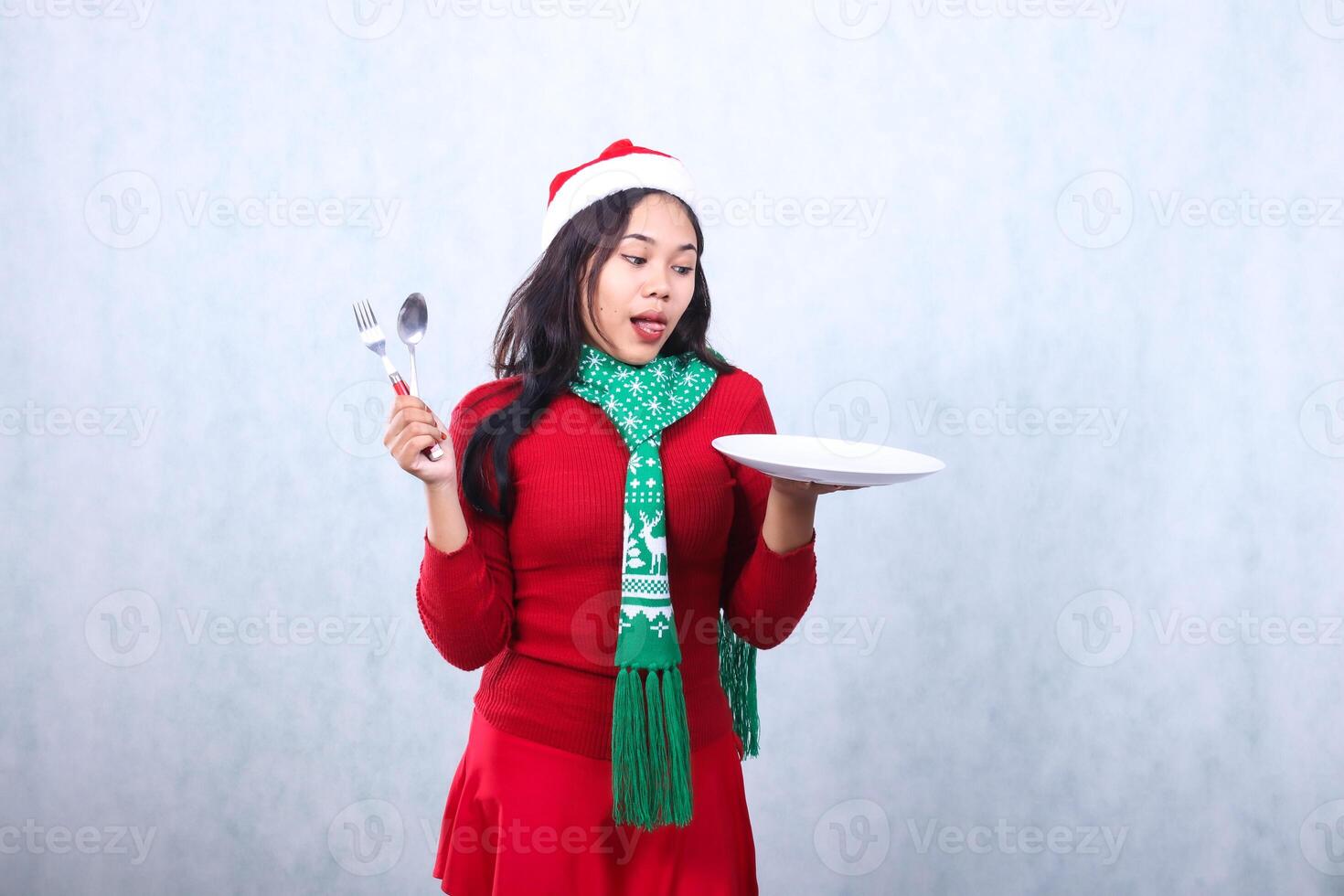 portrait of girl wearing red sweater red sweater with santa hat and scarf celebrating merry christmas , hungry looking at food carry white plate and hold spoon fork ,isolated on white background photo