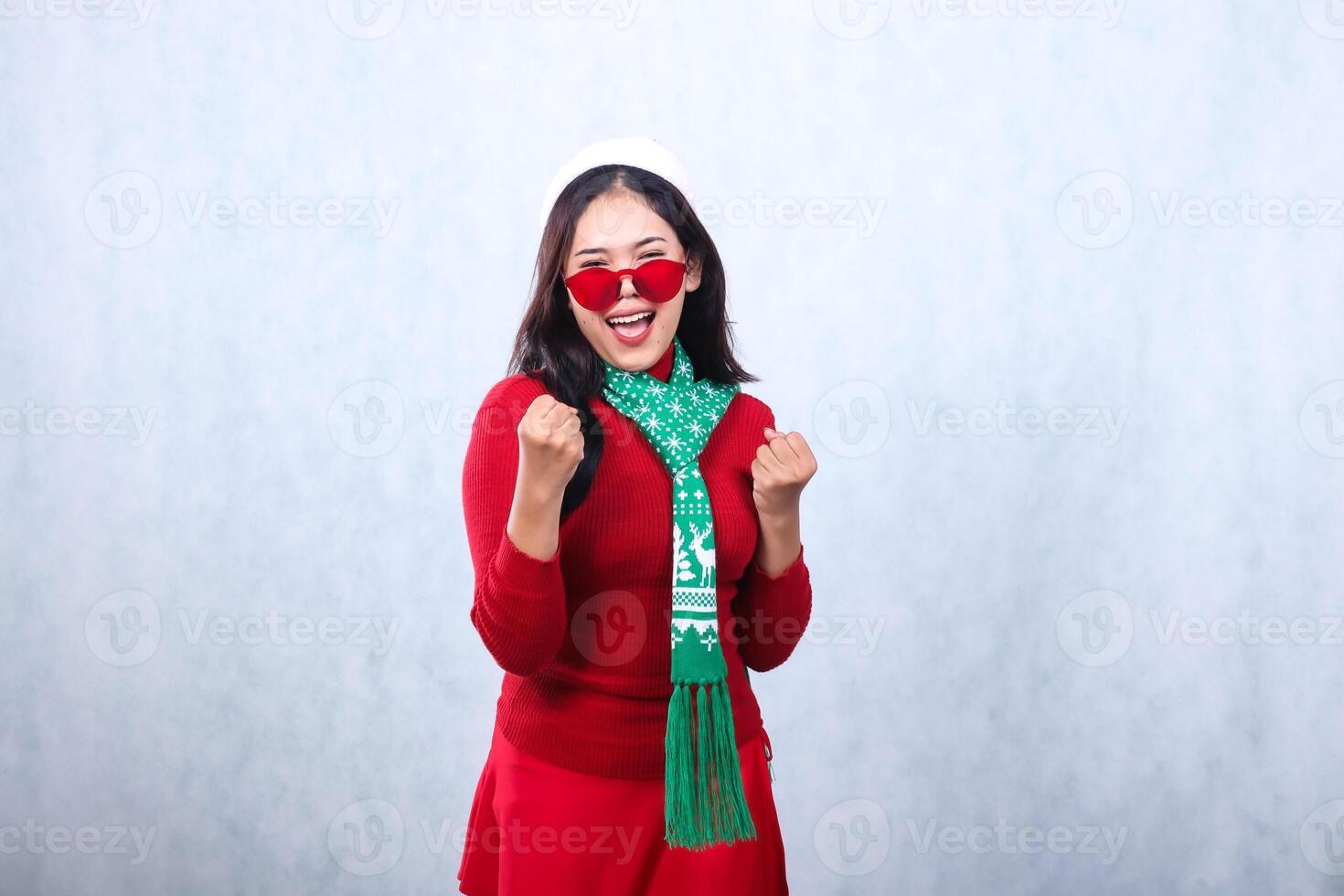 portrait woman wearing red sweater with Santa hat and scarf celebrating merry christmas, cheerful to camera wearing glasses both hands clenched in front of arms, isolated on white background photo
