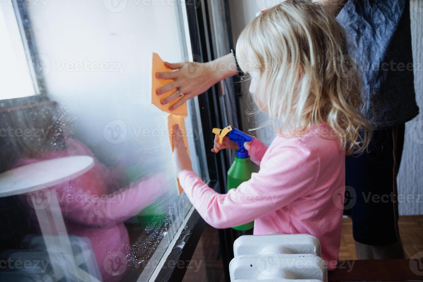 Young child helps clean a window with a parent using a spray bottle and cloth in a bright living room photo