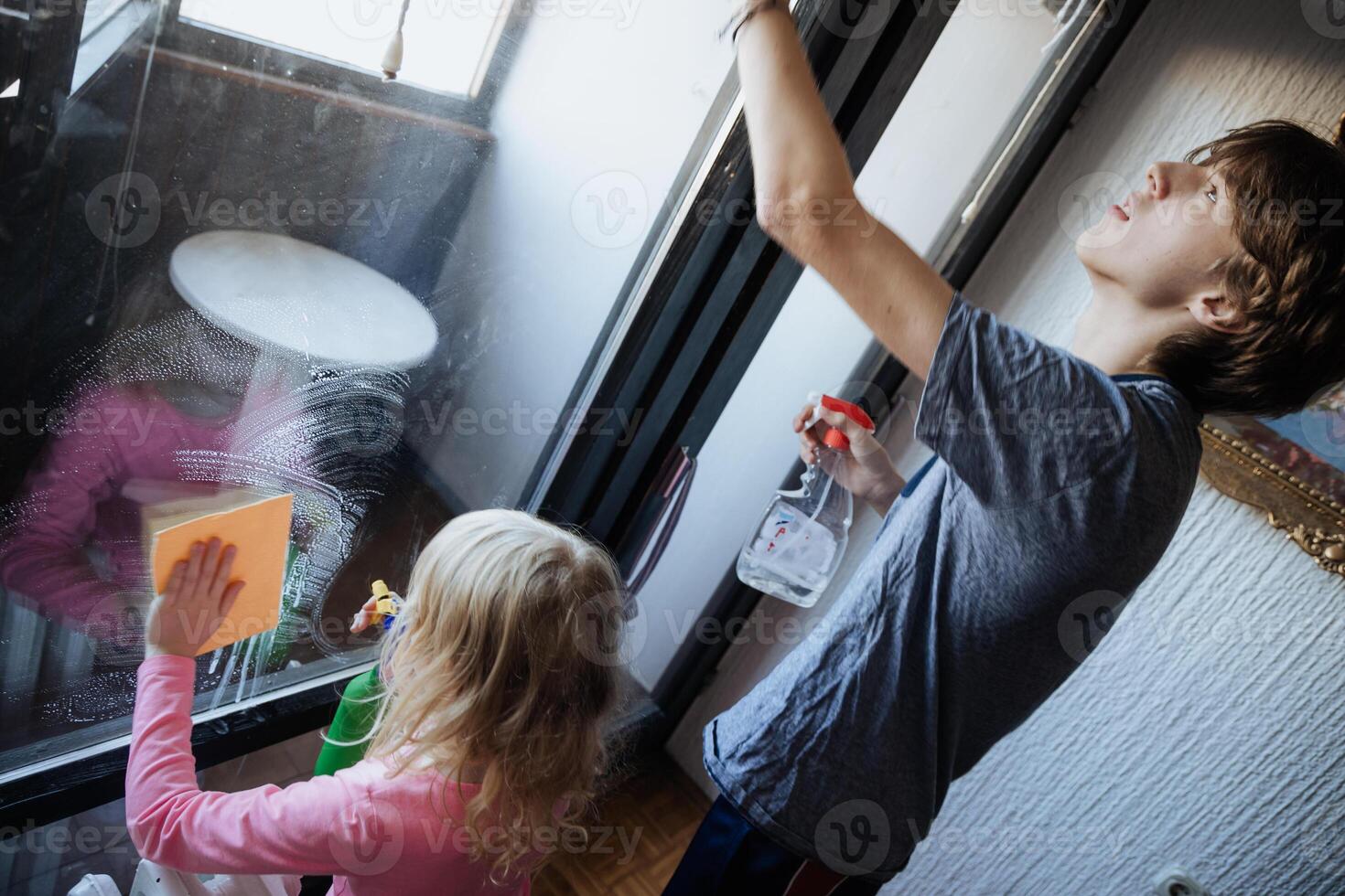 Child cleaning a window while an adult supervises in an indoor setting during a sunny day photo