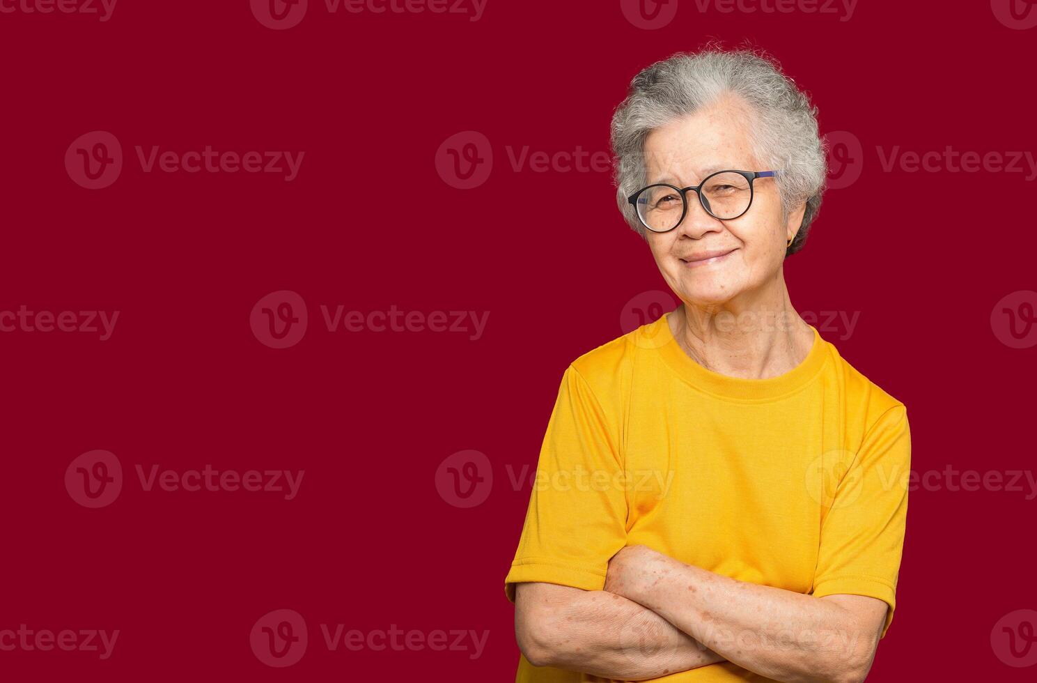 An elderly Asian woman with short gray hair looking at the camera with a smile while standing arms crossed on a red background photo