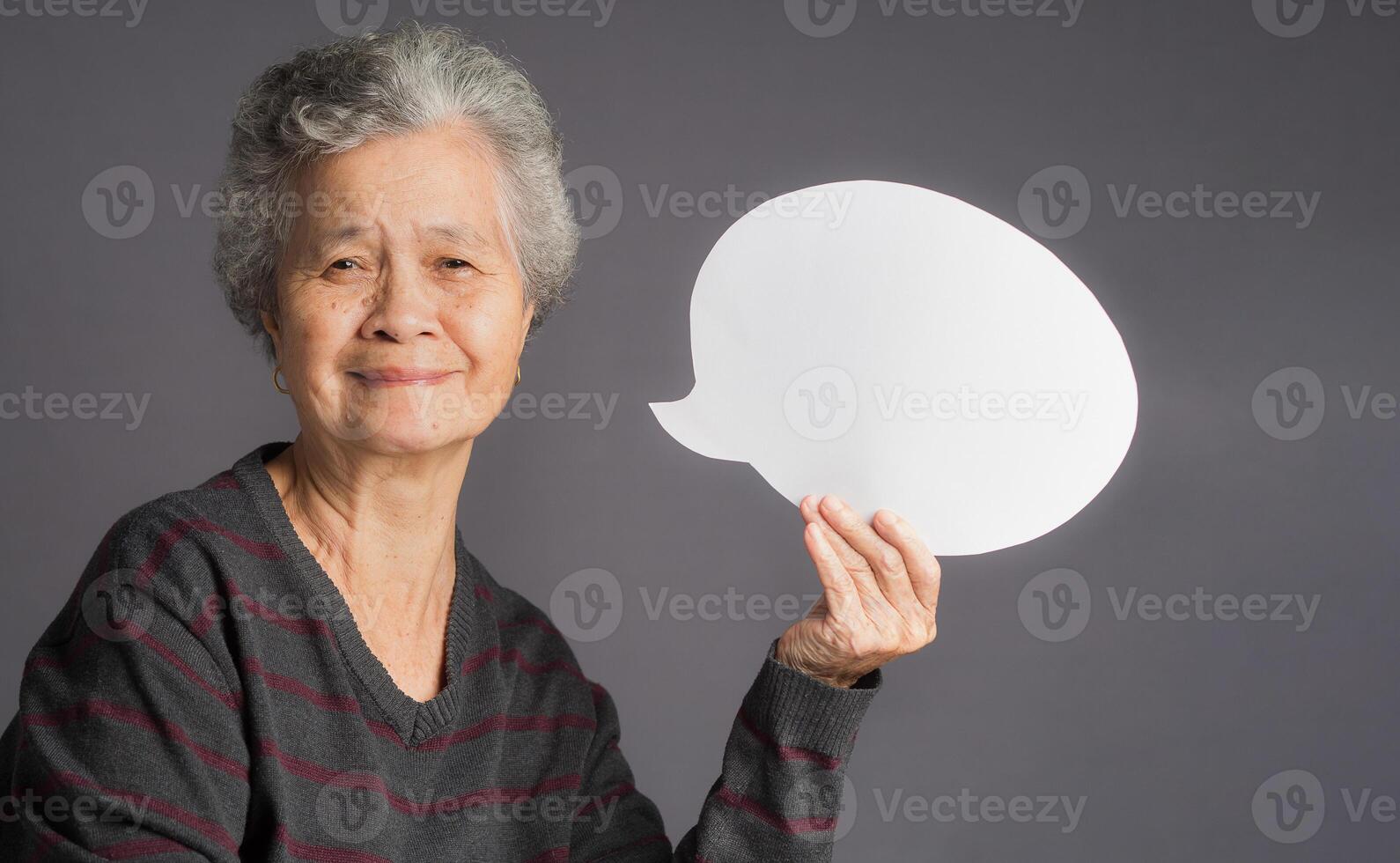 An elderly Asian woman holding a blank speech bubble and looks at the camera with a smile while standing on a gray background photo