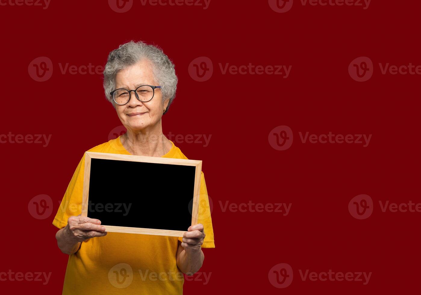 Portrait of an elderly Asian woman with short gray hair holding a mini blackboard and looking at the camera with a smile while standing on a red background photo