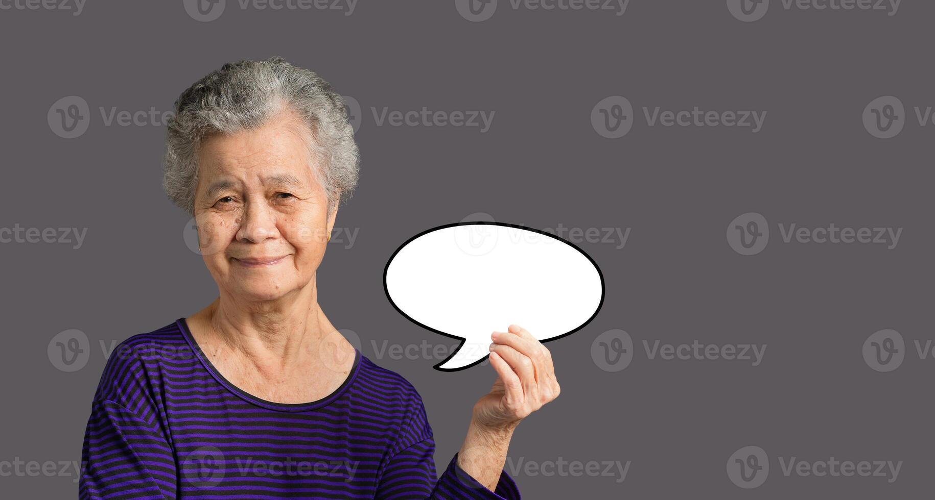 Portrait of an elderly Asian woman is holding a blank speech bubble with a smile and looking at the camera while standing on a gray background photo