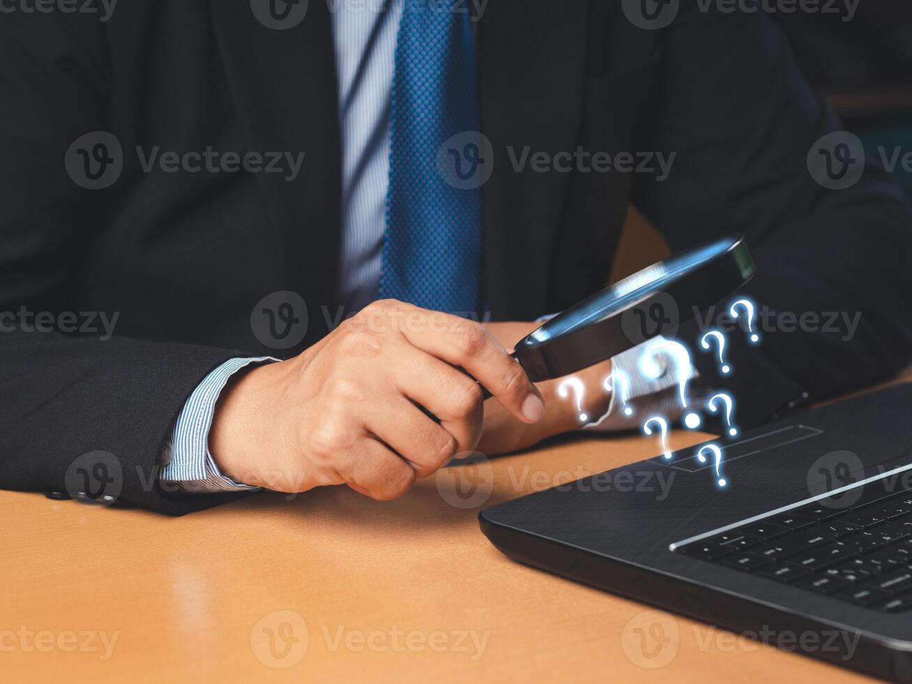 A businessman is holding a magnifying glass and looking at a question mark symbol on a laptop while sitting in the office photo