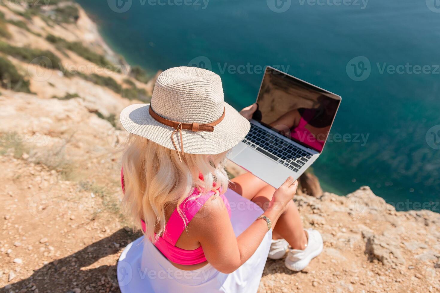 A woman is sitting on a rocky beach with a laptop open in front of her. She is wearing a pink top and a straw hat. Concept of relaxation and leisure. photo