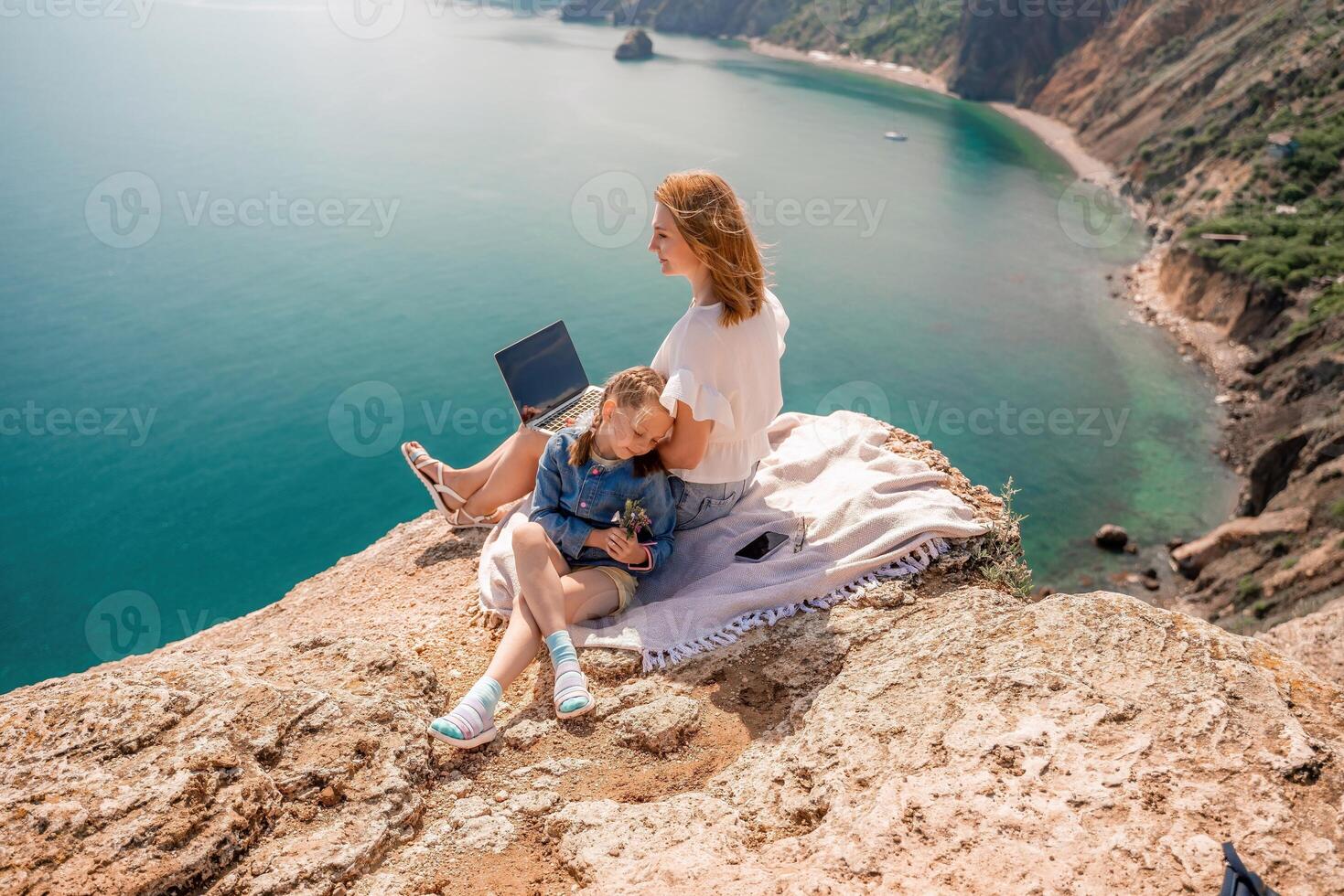Freelance woman with her daughter working on a laptop by the sea, typing on the keyboard, enjoying the beautiful view, highlighting the idea of remote work. photo