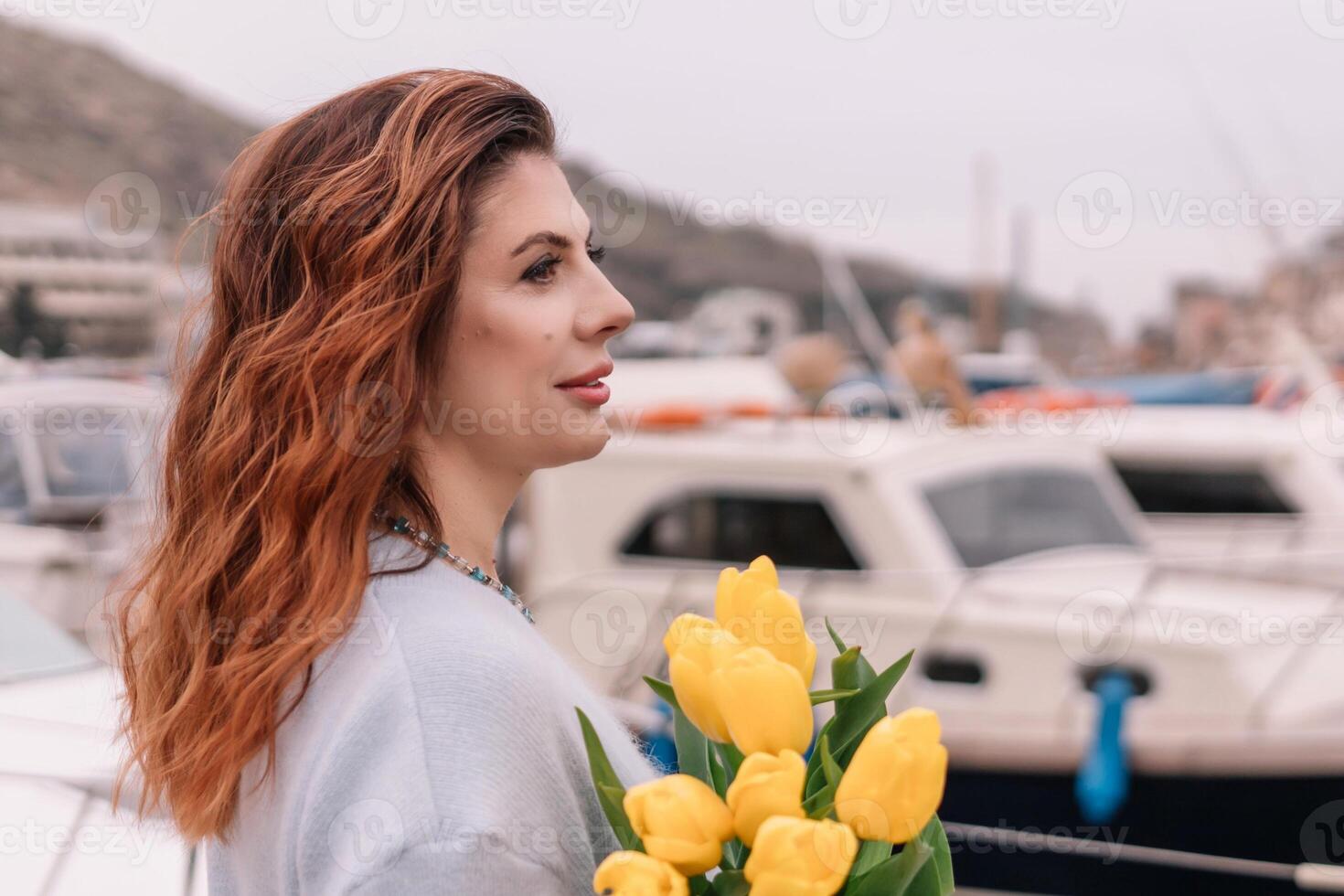 Woman holds yellow tulips in harbor with boats docked in the background., overcast day, yellow sweater, mountains photo