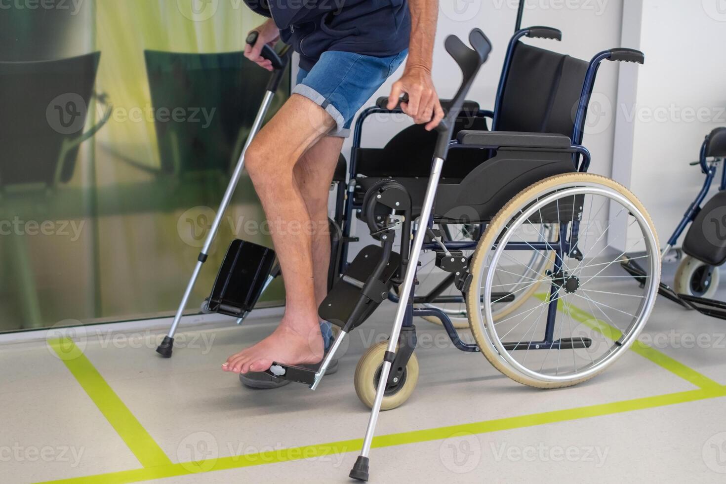 man in shorts on crutches stands next to a wheelchair in a medical facility while waiting to be examined by a doctor, photo