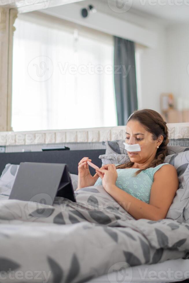 woman lies in bed in casual clothes and with bandages on her face after plastic surgery on her nose, files nails and watches favorite movie on tablet, photo