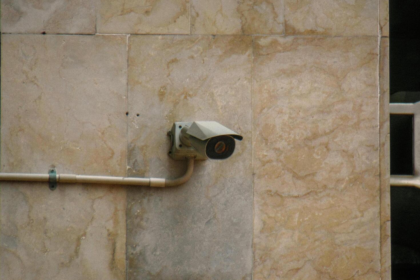 A security camera mounted on a beige marble wall, connected by a conduit cable, symbolizing surveillance, safety, and modern security systems. photo