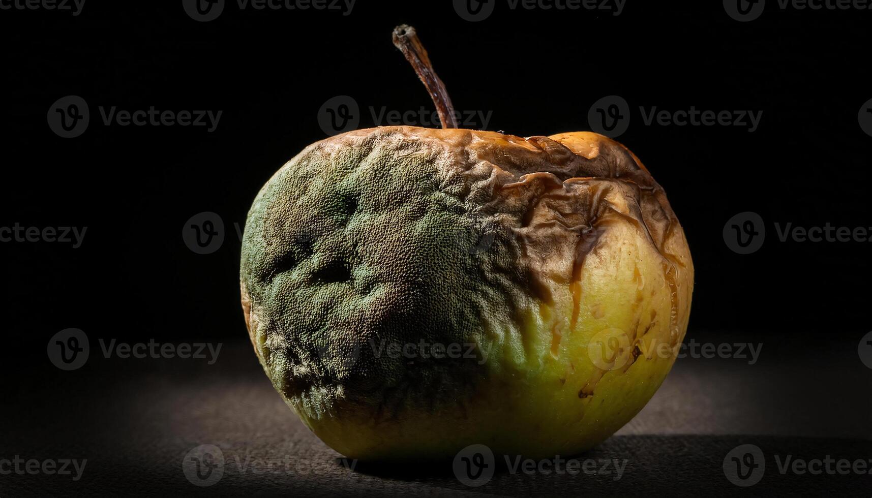 close-up of a rotting apple with brown spots and green mold growth spreading across its wrinkled surface under low light photo