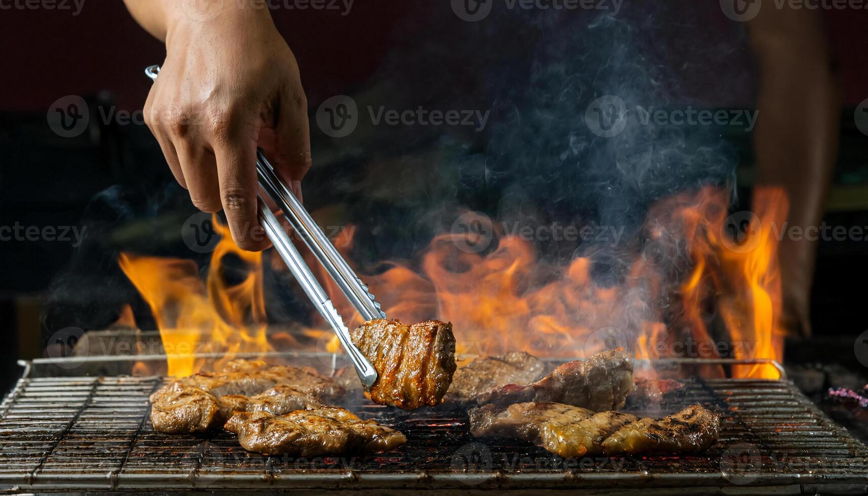 close-up of hands using tongs to grill juicy meat over open flames, with smoke rising on a barbecue grill photo