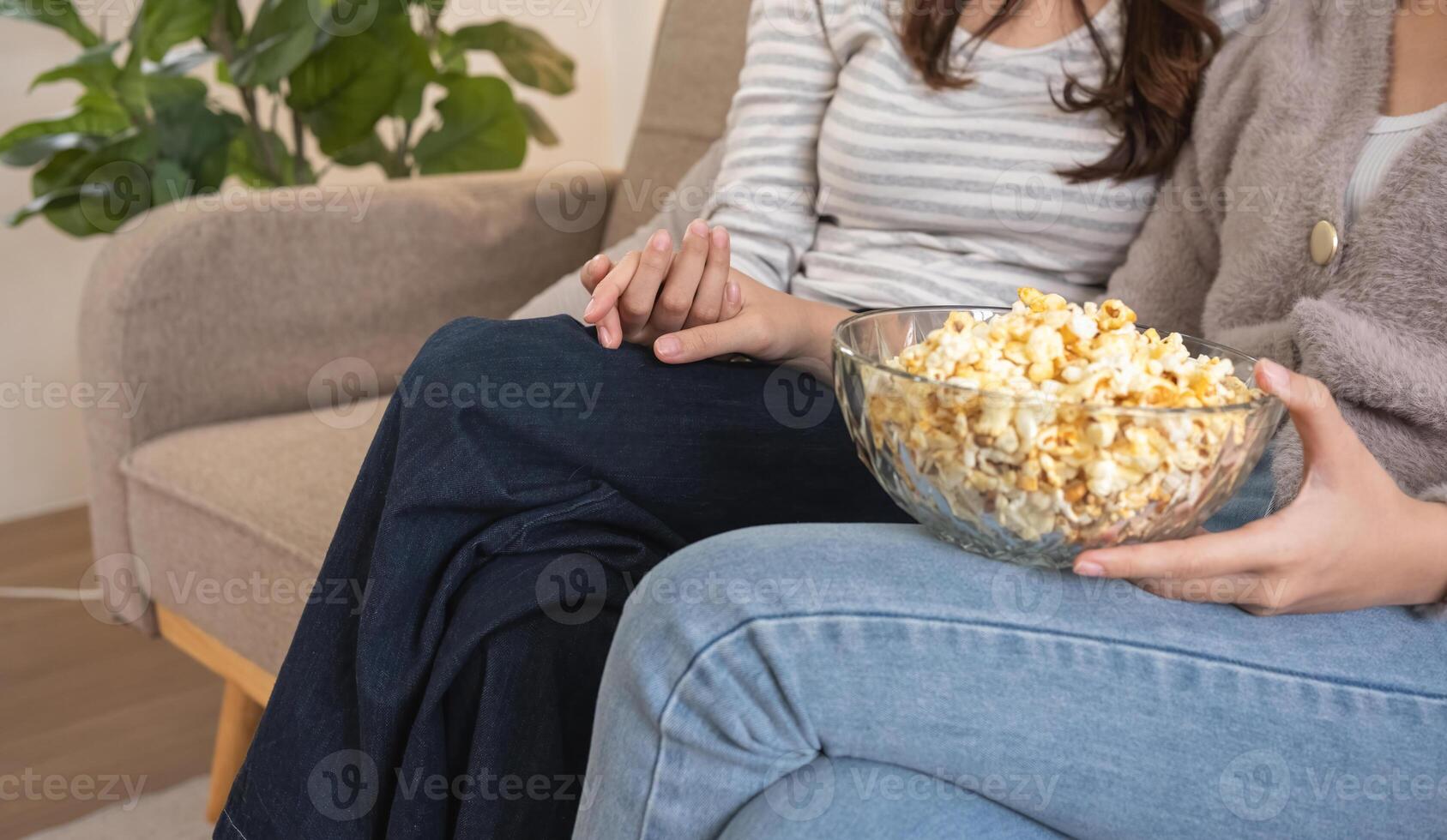 Close-up of two women holding hands with bowl of popcorn on couch. Concept of friendship and comfort photo