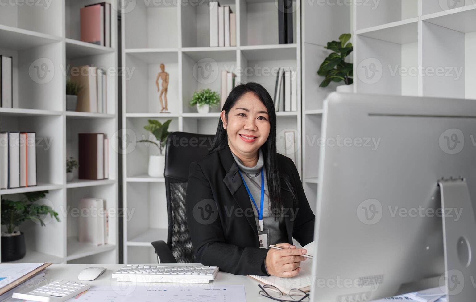 Middle-aged Business Woman Sitting in Modern Office, Smiling at Desk with Computer, Surrounded by Bookshelves and Office Supplies photo