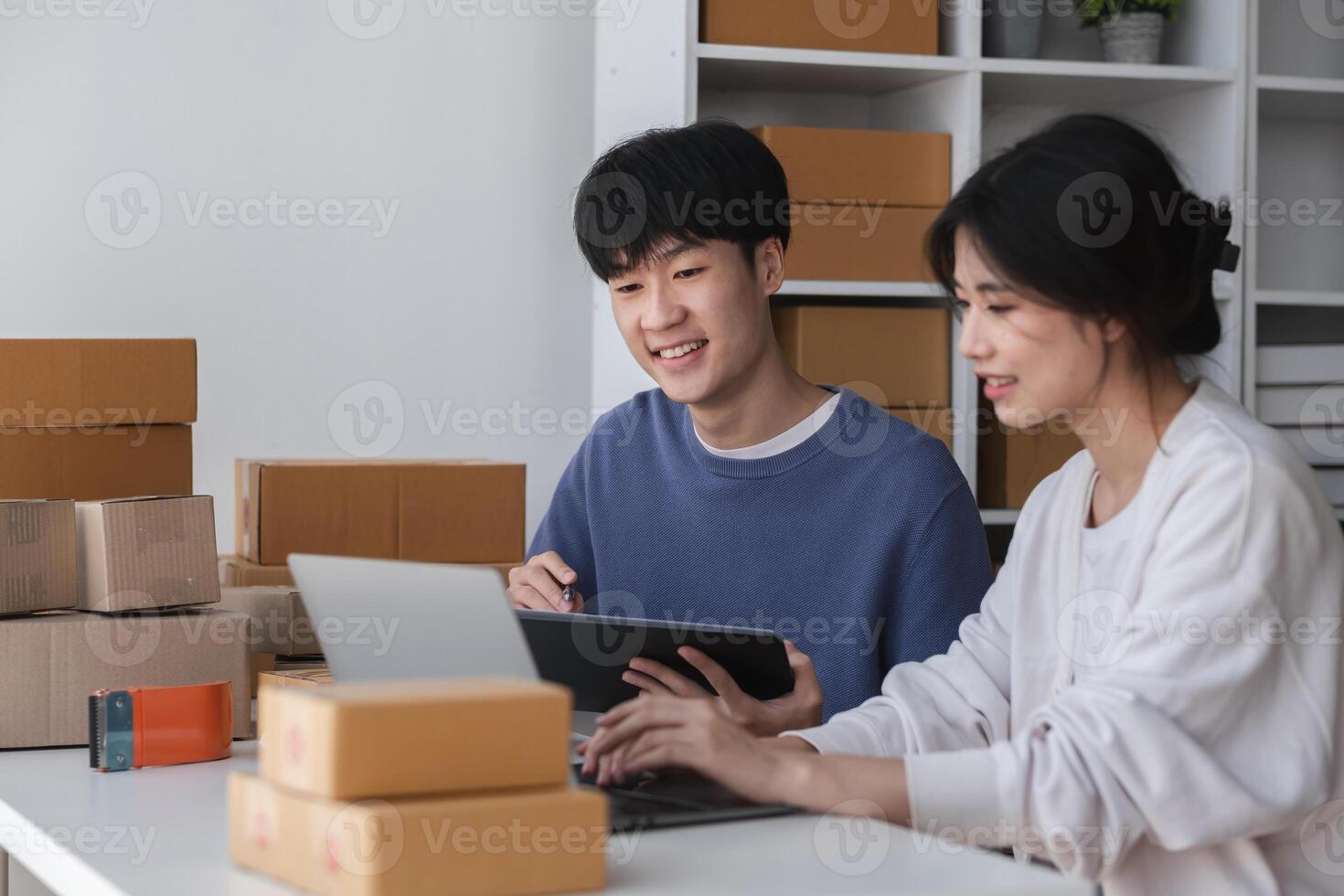 Young Couple Managing Their Online Small Business from Home, Surrounded by Shipping Boxes and Working on a Laptop and Tablet in a Modern Home Office Setting photo