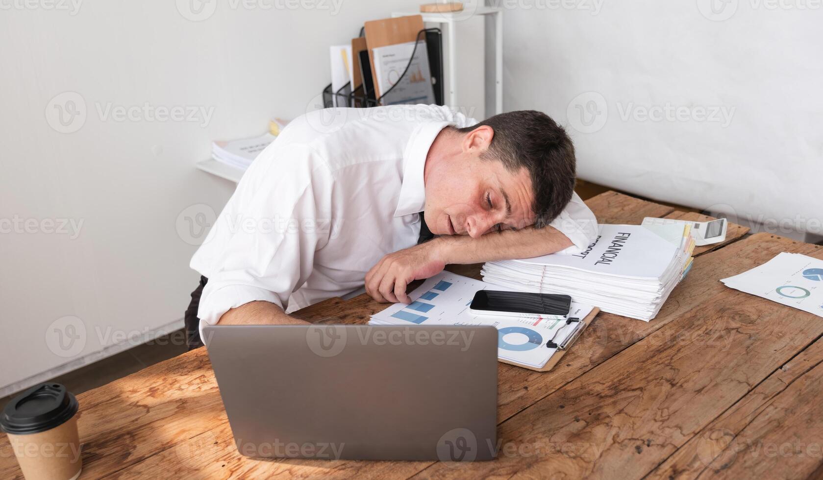 Businessman exhausted and asleep at desk with laptop and financial reports, concept of workplace fatigue and stress photo