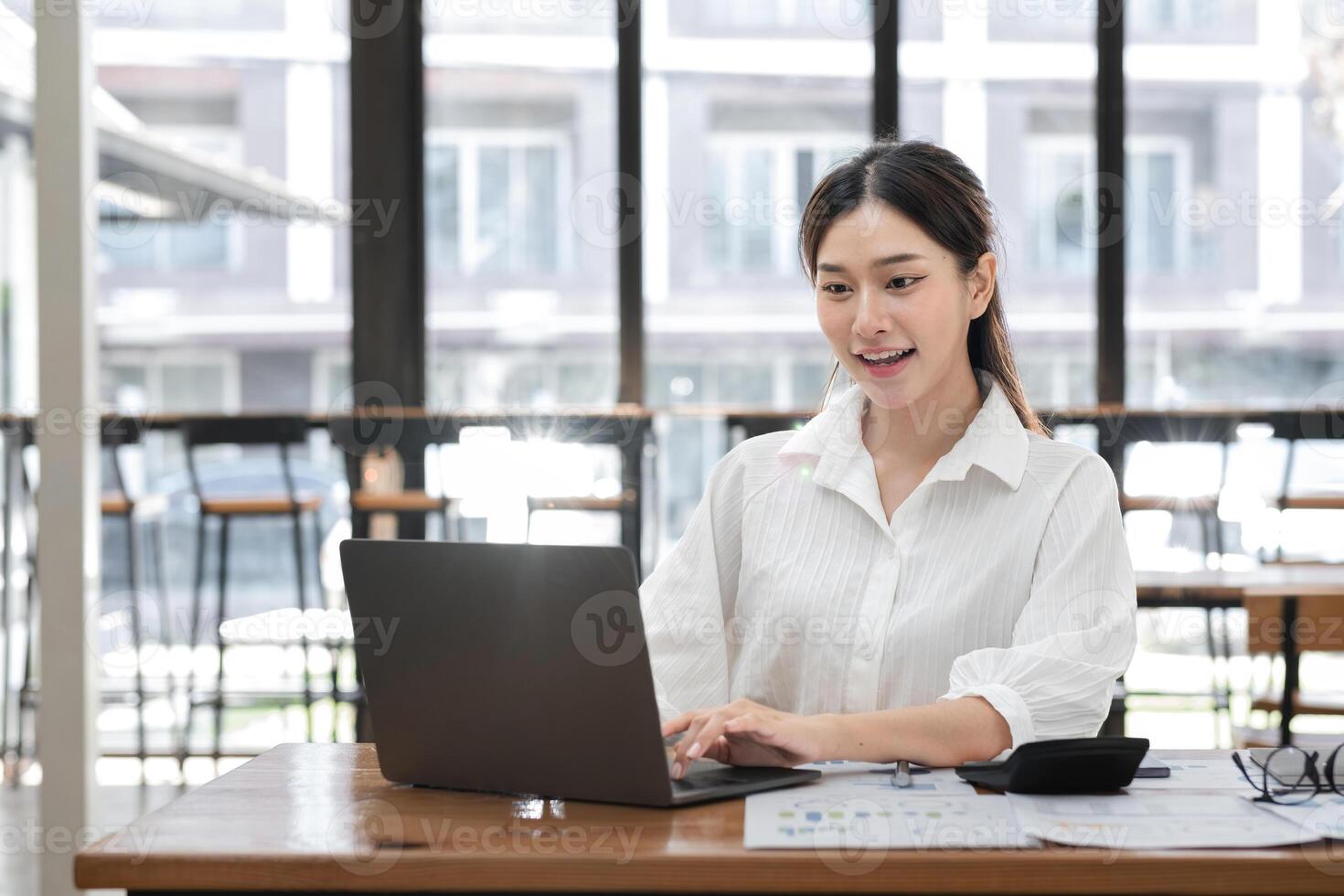 Professional Business Woman Working on Laptop in Modern Office Setting with Documents and Calculator on Desk, Smiling and Engaged in Productive Work, Large Windows in Background photo