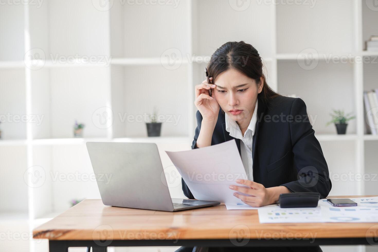 Stressed Business Woman Working at Desk with Laptop and Documents in Modern Office Environment, Experiencing Workload Pressure and Deadlines, Wearing Formal Attire, Concentrating on Paperwork photo