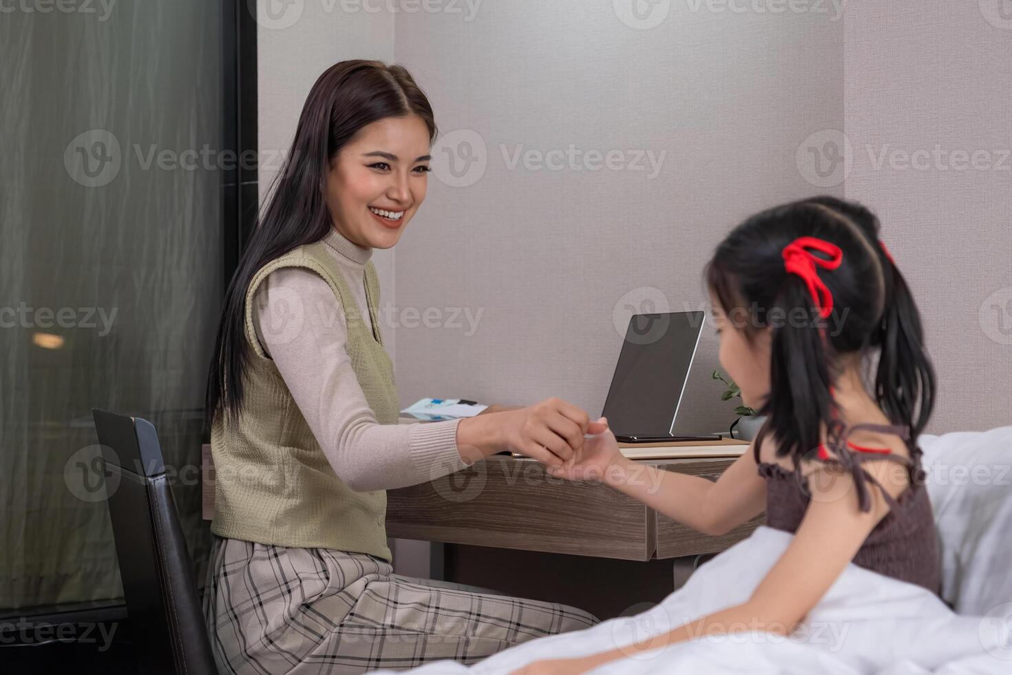Woman Working on Laptop and Holding Hands with Daughter in Bedroom. Concept of work-life balance and family bonding. photo