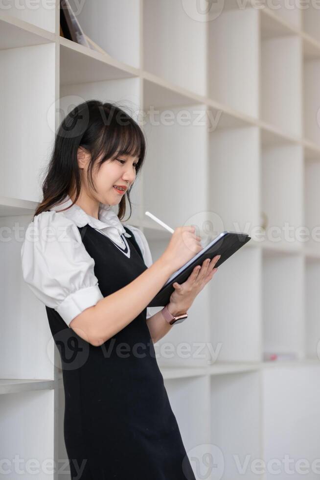 Young Female University Student Taking Notes on Digital Tablet in Modern Library Setting, Focused on Learning and Education, Wearing Casual Outfit, Engaged in Academic Activities, Smiling photo