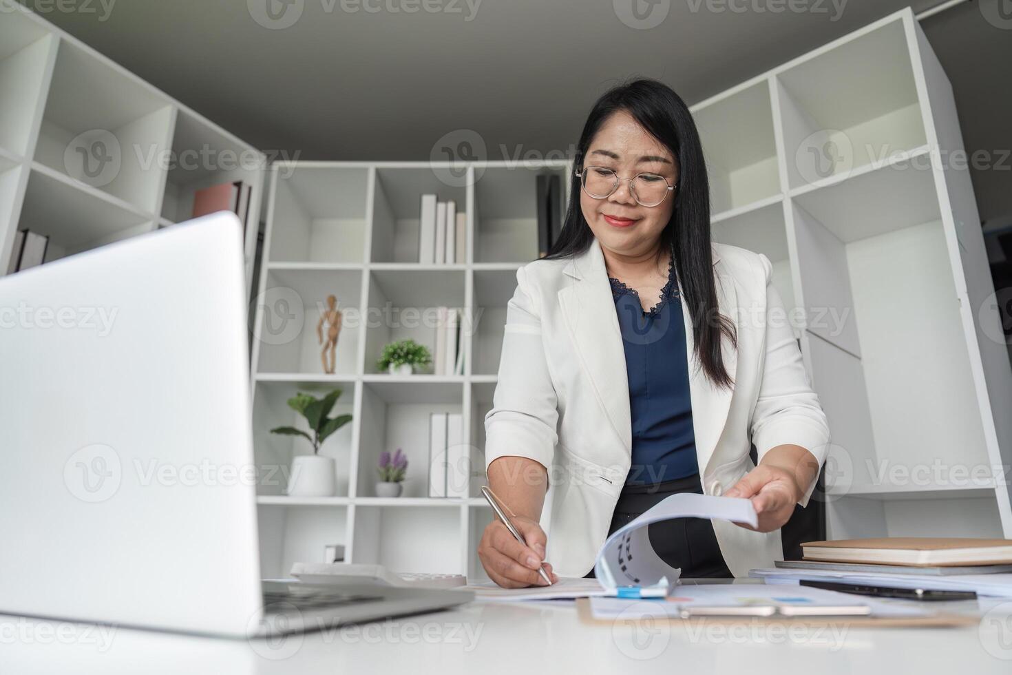 Middle-aged Business Woman Doing Paperwork at Desk in Modern Office with Computer and Shelves in Background photo