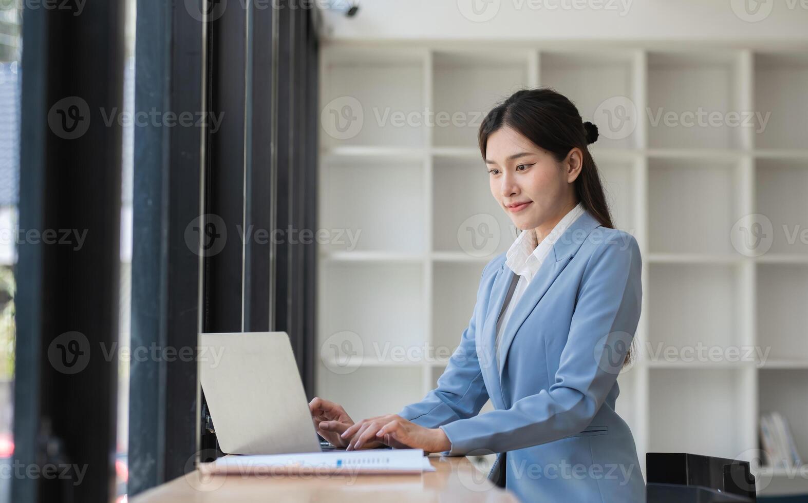 Professional Business Woman Working on Laptop in Modern Office Setting with Natural Light and Minimalist Design, Focused and Productive, Wearing Formal Attire, Engaged in Corporate Tasks photo
