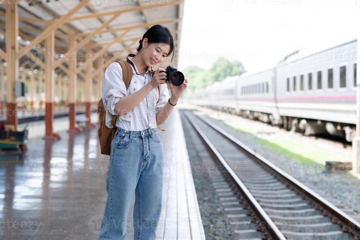 Young Asian woman holds a camera and takes photos of a train while waiting for the train at the train station to travel.