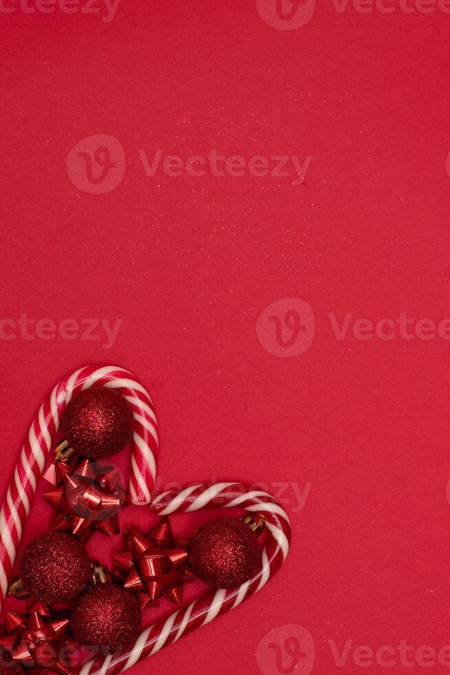 Top view photo heart lined with Christmas lollipops and decorated with Christmas tree toys of red colour,on red background for greeting card. Xmas backdrop with space for text.Copyspace.Vertical photo.