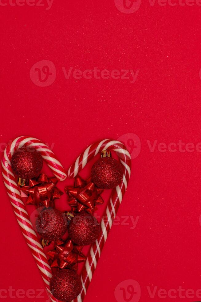 Top view photo heart lined with Christmas lollipops and decorated with Christmas tree toys of red colour,on red background for greeting card. Xmas backdrop with space for text.Copyspace.Vertical photo.