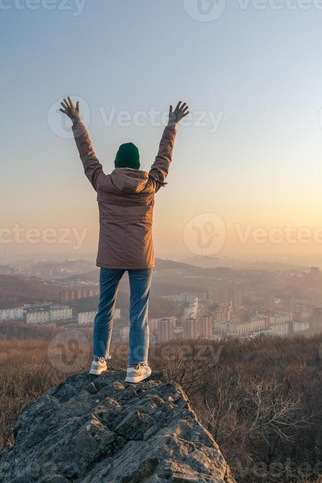 A girl in a jacket and a hat stands on a rock and looks at the city from a height during sunset photo