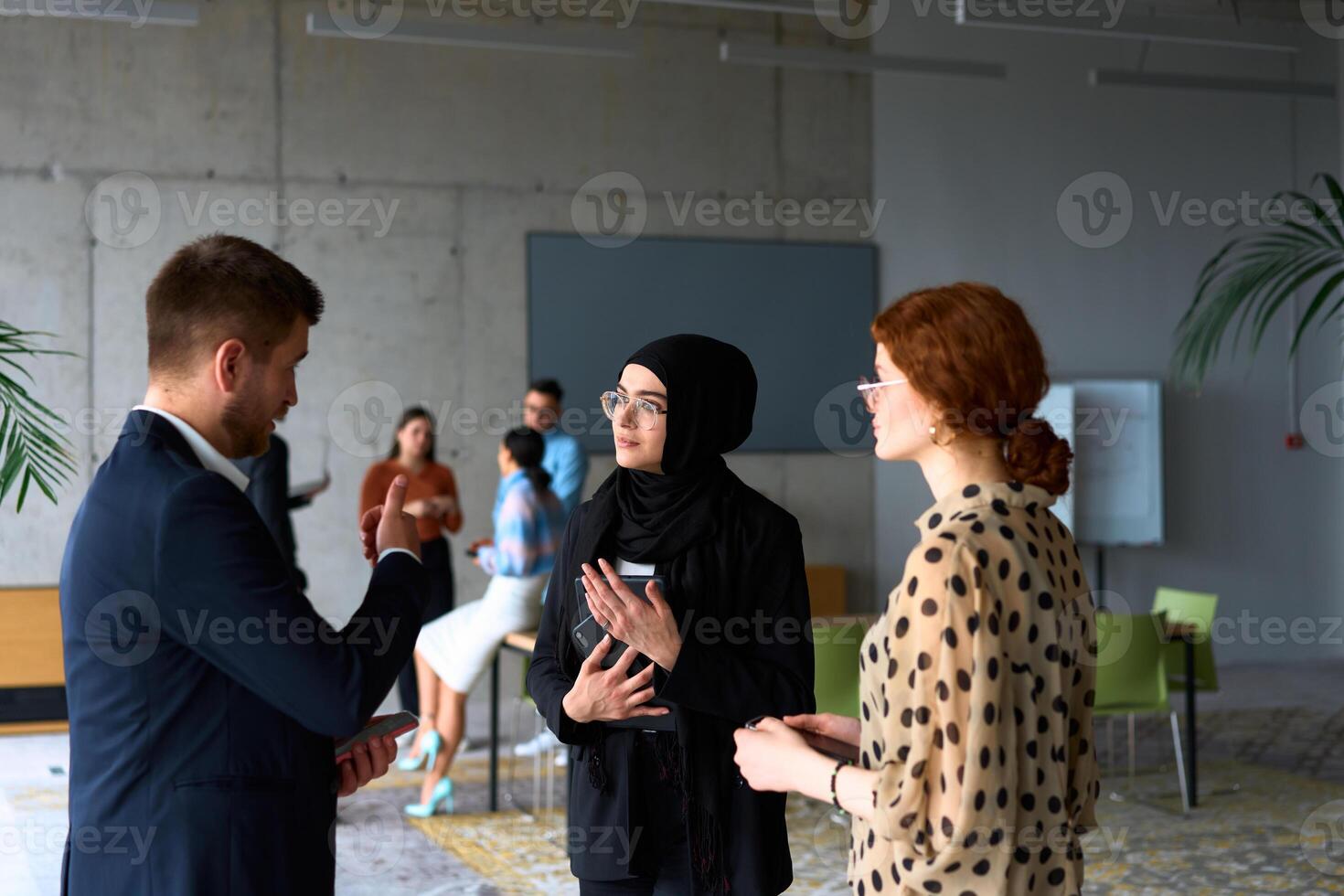 A diverse group of business professionals engages in a conversation during a work break in the office, with their colleagues visible in the background, fostering collaboration and connection photo