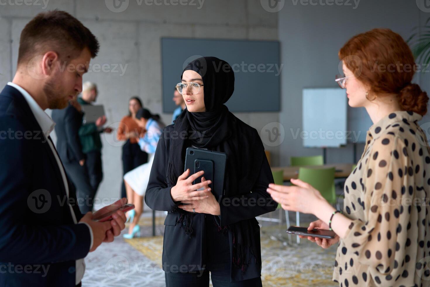 A diverse group of business professionals engages in a conversation during a work break in the office, with their colleagues visible in the background, fostering collaboration and connection photo
